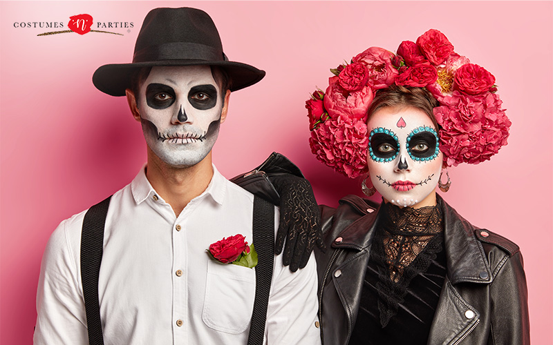 Two people in Day of the Dead costumes with skull makeup pose against a pink background.